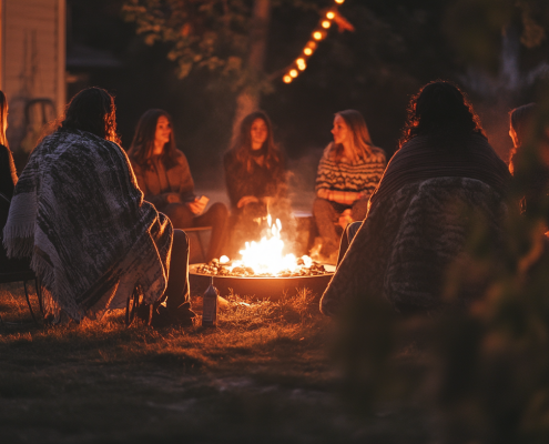 Group of women sitting quietly around a glowing campfire in nature, wrapped in blankets, sharing warmth and stillness under the evening sky.